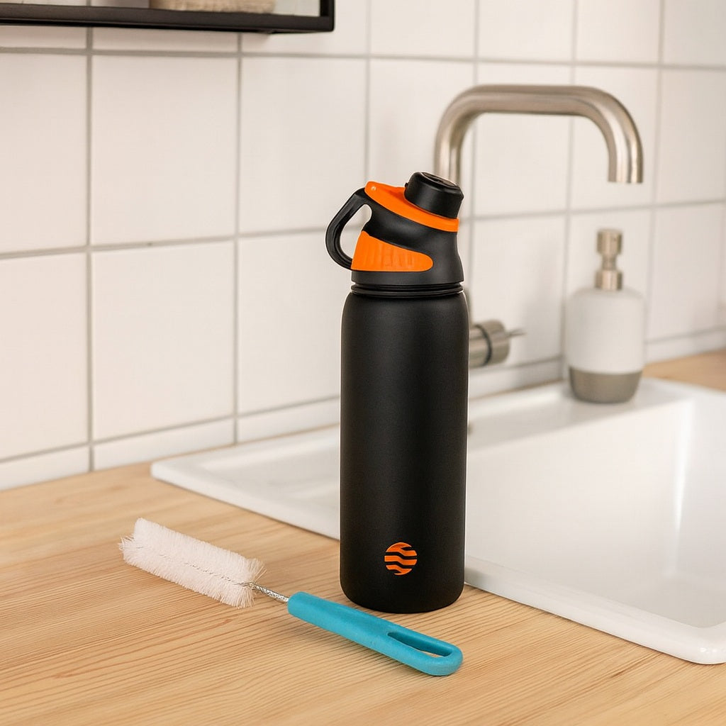 Gourde isotherme noire avec bouchon sport orange posée sur un plan de travail près d’un évier, accompagnée d’une brosse de nettoyage.