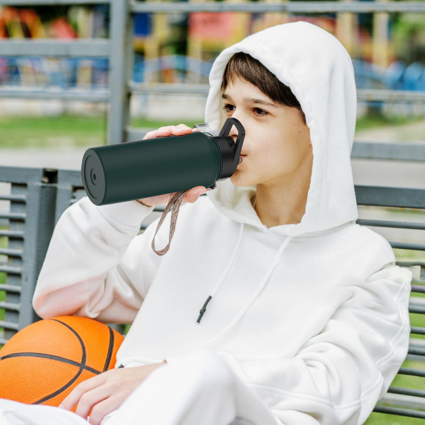 Adolescent en hoodie blanc buvant avec une gourde isotherme verte, assis sur un banc avec un ballon de basket